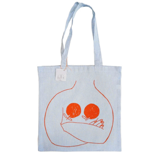 totebag de color vaquero claro con una ilustración de una persona cruzada de brazos con dos naranjas en el pecho