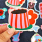 mano sujetando una pegatina de Laura Ortiz en forma de taza de café a rayas rosas y rojas, con el asa en forma de pétalos de flor rojos y un platito verde