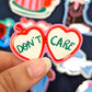 mano sujetando una pegatina de Laura Ortiz en forma de colgante con forma de corazón rojo abierto y el texto DON'T CARE en color verde