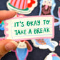 mano sujetando una pegatina de Laura Ortiz en forma de un trozo de libreta con el texto It's okay to take a break