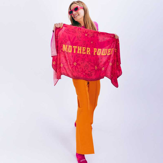 Mujer vestida de rosa y naranja con gafas de sol susteniendo un pañuelo de cuello rosa con el mensaje "Mother power"