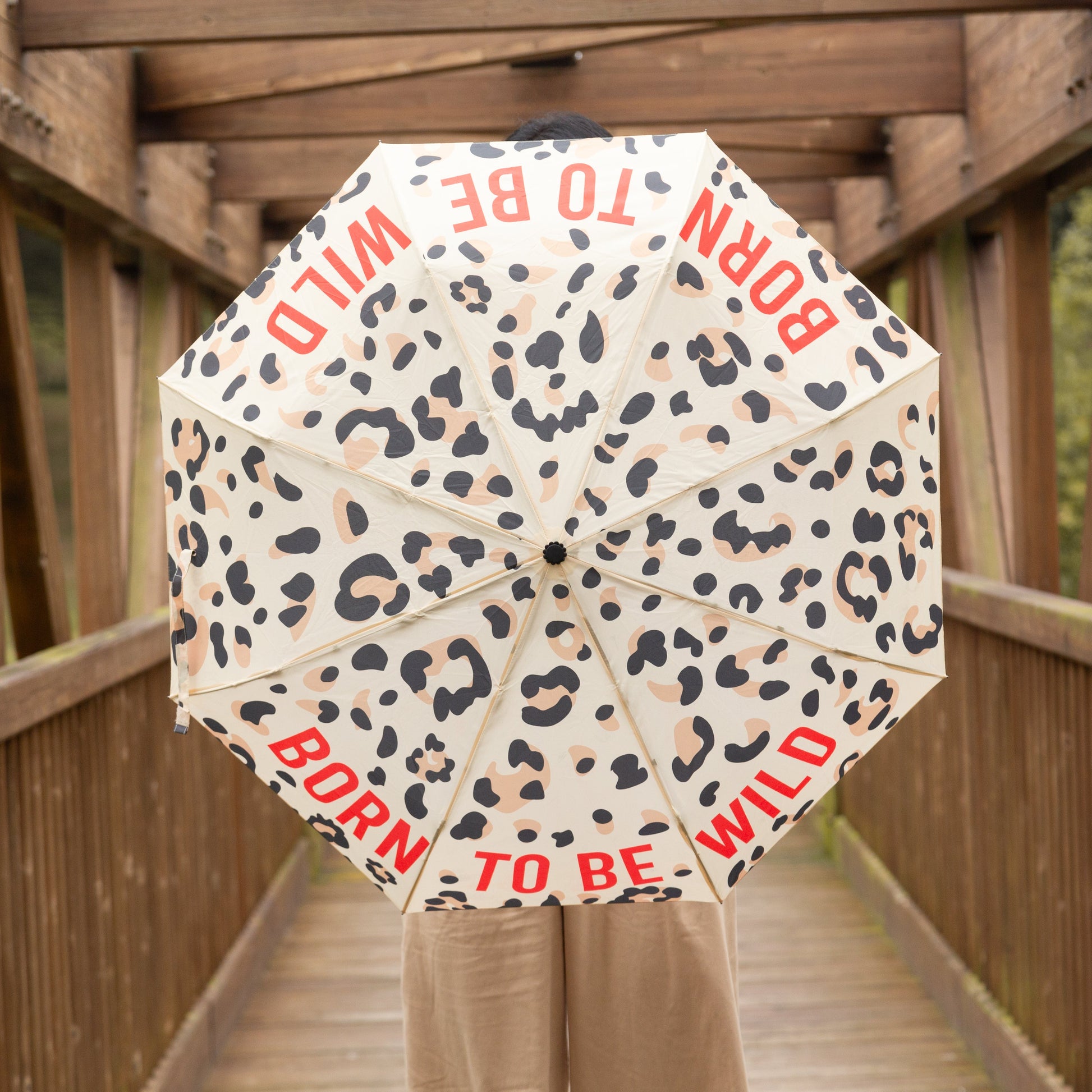 Mujer en un puente de madera con un paraguas plegable con esqueleto de fibra de vidrio negro, estampado de leopardo y el mensaje Born to be wild escrito en letras mayúsculas rojas