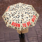 Mujer bajo la lluvia con un paraguas plegable con esqueleto de fibra de vidrio negro, estampado de leopardo y el mensaje Born to be wild escrito en letras mayúsculas rojas