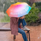 Chica bajo la lluvia con un paraguas plegable con esqueleto negro y estampado degradado de colores con el mensaje Hello rain, welcome back en letras mayúsculas blancas