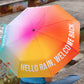 Chica sentada de espaldas con el paraguas plegable con esqueleto negro y estampado degradado de colores con el mensaje Hello rain, welcome back en letras mayúsculas blancas