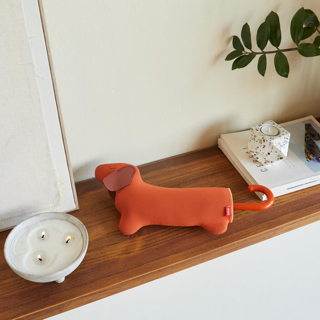 Paraguas plegable Balvi en su funda con forma de perro salchicha marrón, sobre una repisa de madera.
