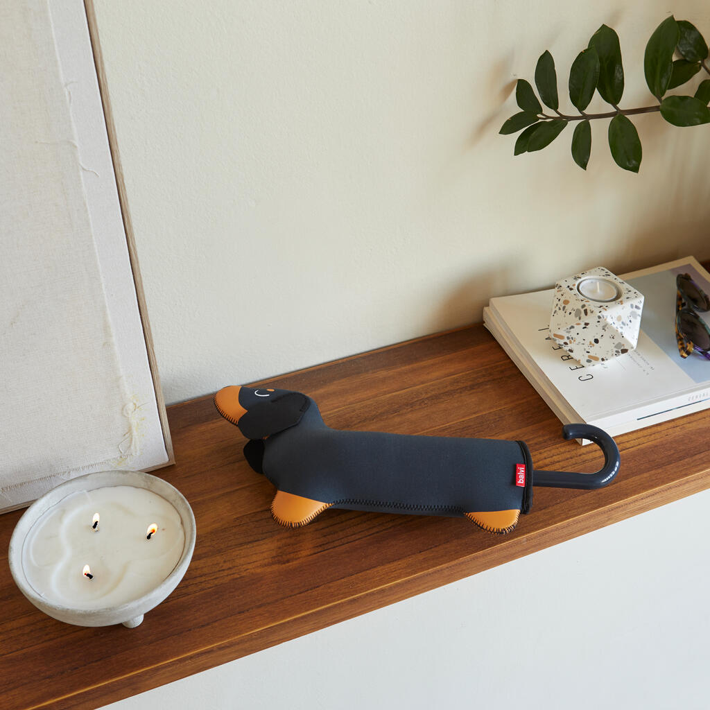 Paraguas plegable en su funda con forma de perro salchicha negro y marrón, sobre una repisa de madera.