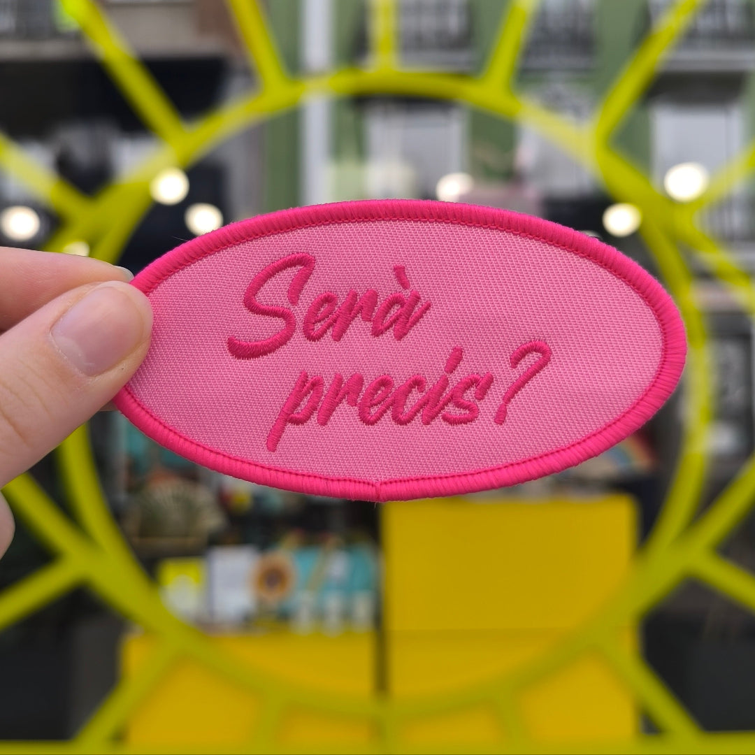 PArche en color rosa con la frase "Serà Precís" sujetado por una mano. 