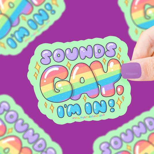 Pegatina de fondo verde con el texto "Sounds Gay I'm In!" con la palabra gay en arcoíris y chispas alrededor sujetado por una mano. 
