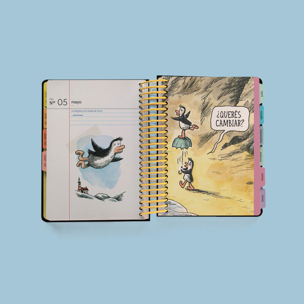 Interior de la Agenda diaria 2027 Macanudo abierta en mayo con viñeta de pingüino volando y página ilustrada a color con la pregunta ¿Querés cambiar?
