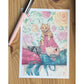 Postal con ilustración de Esther Gili de una chica tumbada con dos gatos encima y una pared con papel pintado de rosas de fondo
