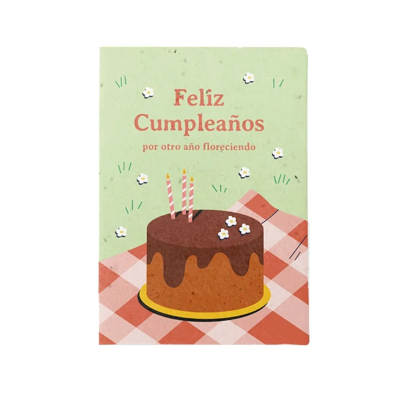 Postal cde cumpleaños con una tarta de chocolate con velas sobre un mantel de cuadros rojos en el césped y el mensaje Feliz Cumpleaños por otro año floreciendo hecha con papel de semillas