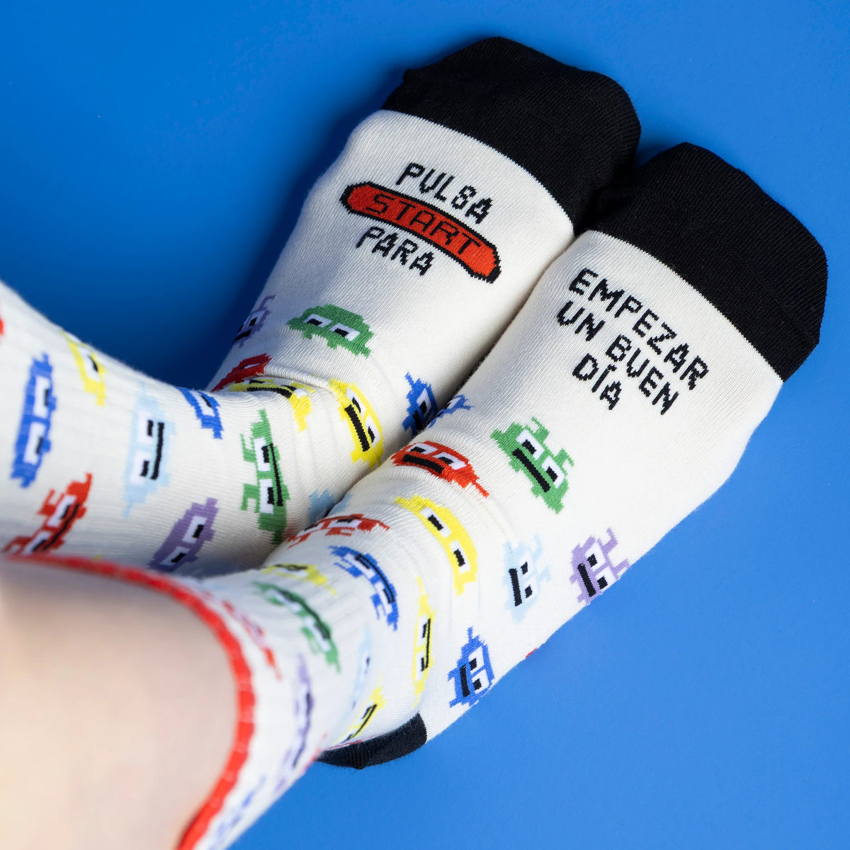 Calcetines UO blancos con personajes de videojuego y mensaje «Pulsa Start para empezar un buen día», puestos sobre fondo azul.