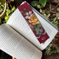 Libro abierto sobre unas plantas con un punto de libro granate con ilustración de Laura Agustí de flores y una liebre saltando