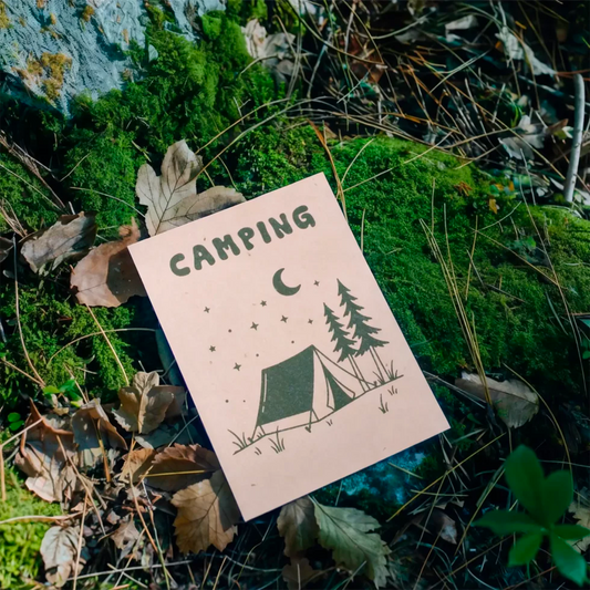 postal con semillas dentro de la marca resetea con un dibujo de una tienda de campaña, unos árboles y la luna y las estrellas con el texto Camping, colocada sobre el suelo de un bosque