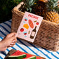 postal con semillas dentro de la marca resetea con dibujos de una barra de pan, dos gildas tomate, queso, copas de vino y una botella de vino y el texto PICNIC, con una cesta con fruta y una toalla de picnic detrás