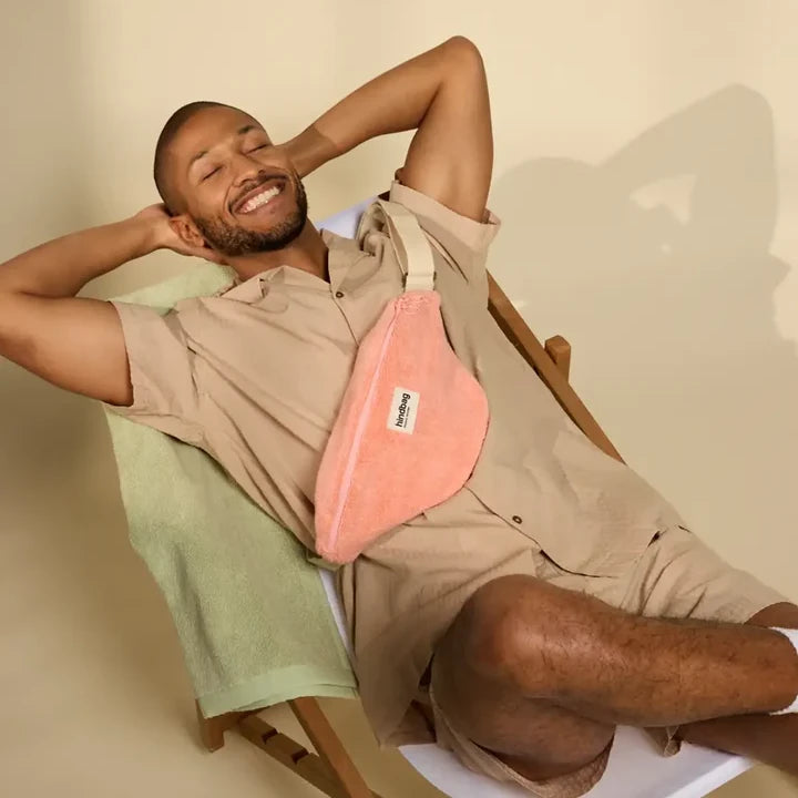 Hombre vestido de beige con una riñonera de rizo rosa de la marca francesa Hindbag sentado en una tumbona sobre toallas del mismo material