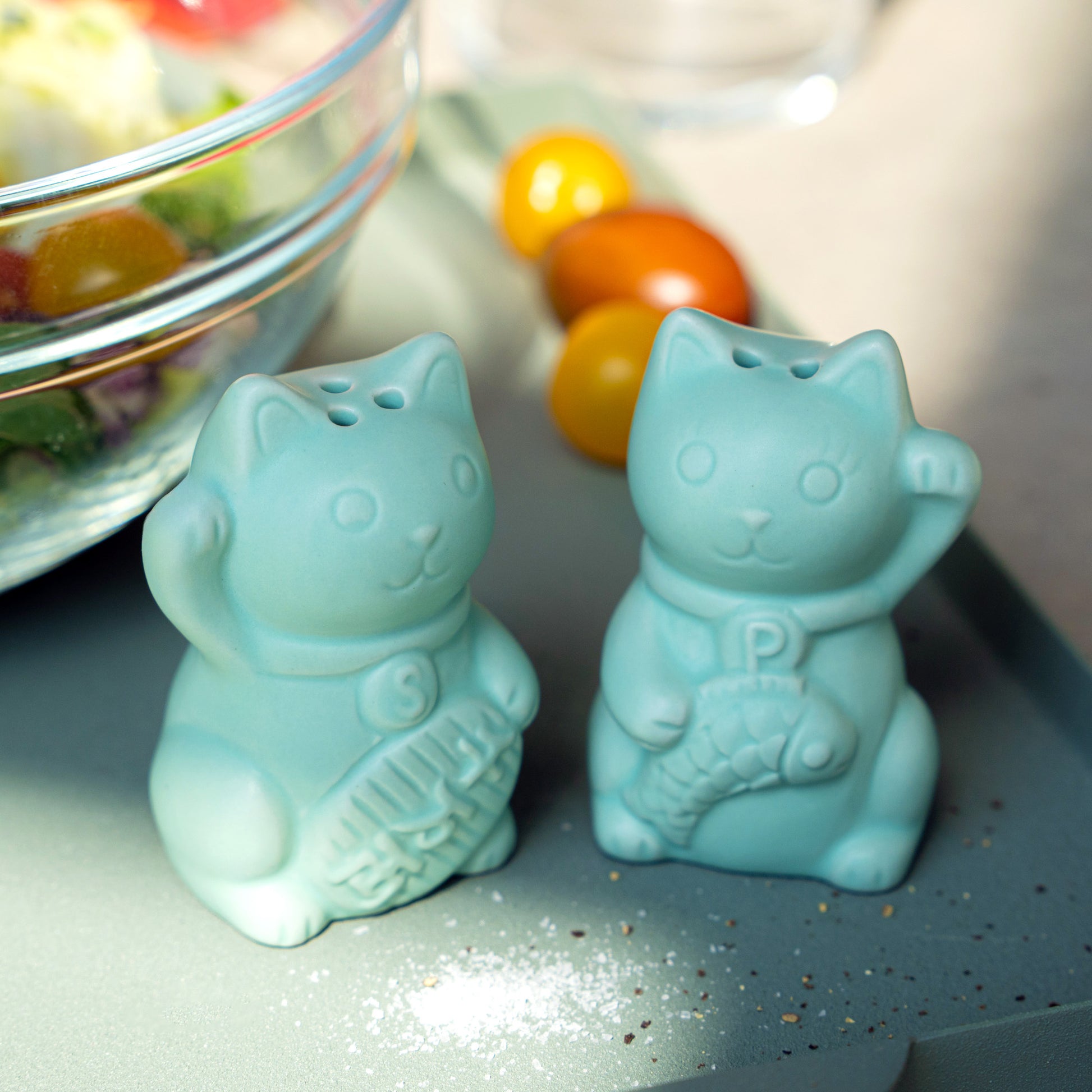Set de sal y pimienta con forma de gato de la suerte verde jade junto a una ensalada de tomates cherry