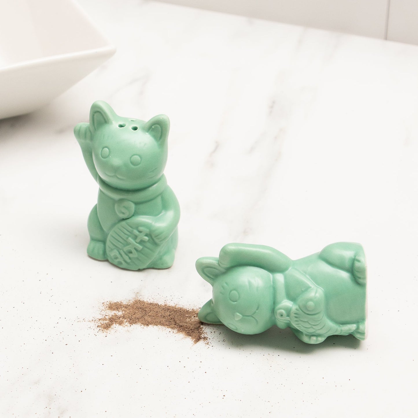 Set de sal y pimienta con forma de gato de la suerte verde jade con el pimentero tumbado y pimienta en la mesa de mármol