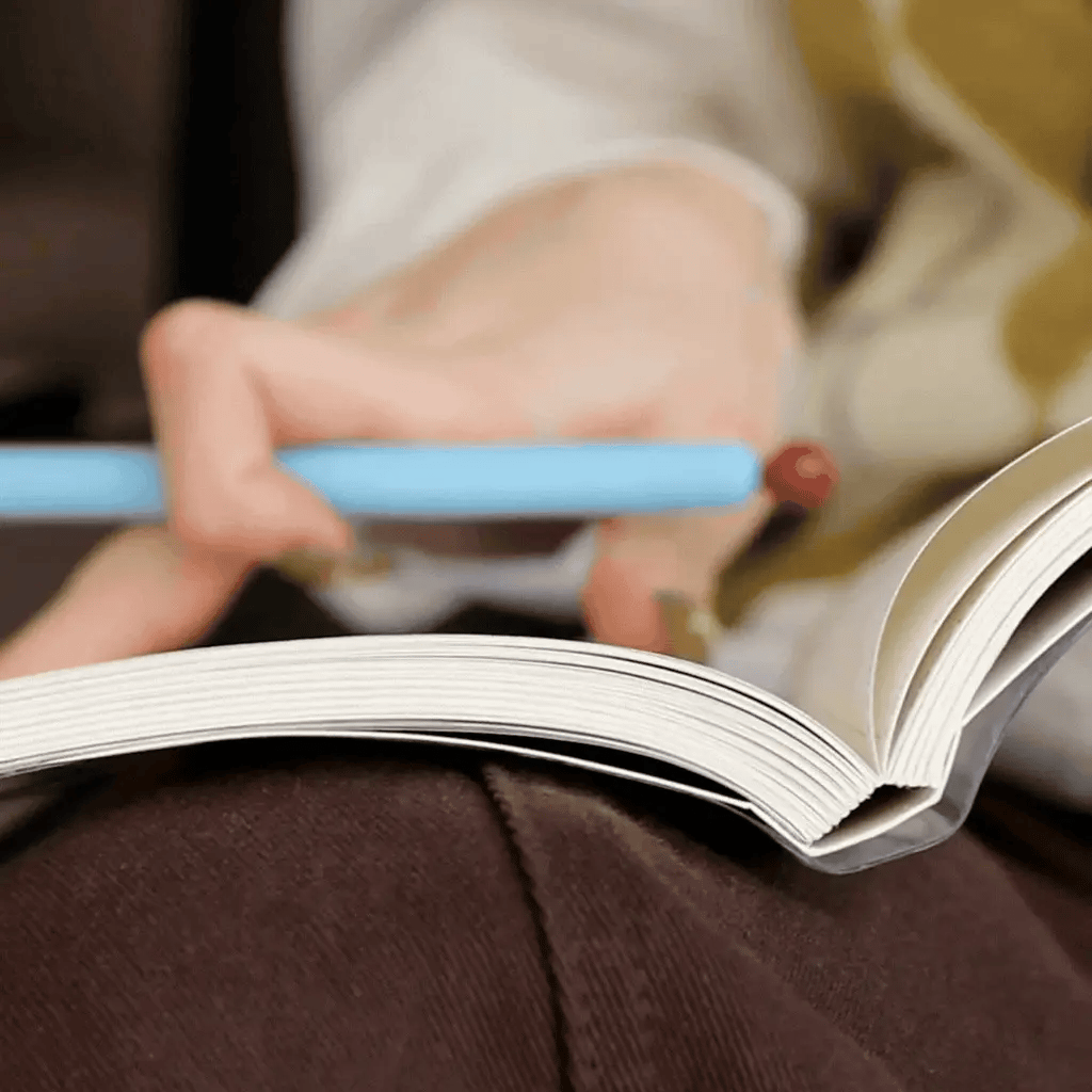 Persona escribiendo en la Agenda 2027 negra Journal Sessions abierta sobre el regazo, con lápiz azul en mano, mostrando el grosor de las 224 páginas cosidas

