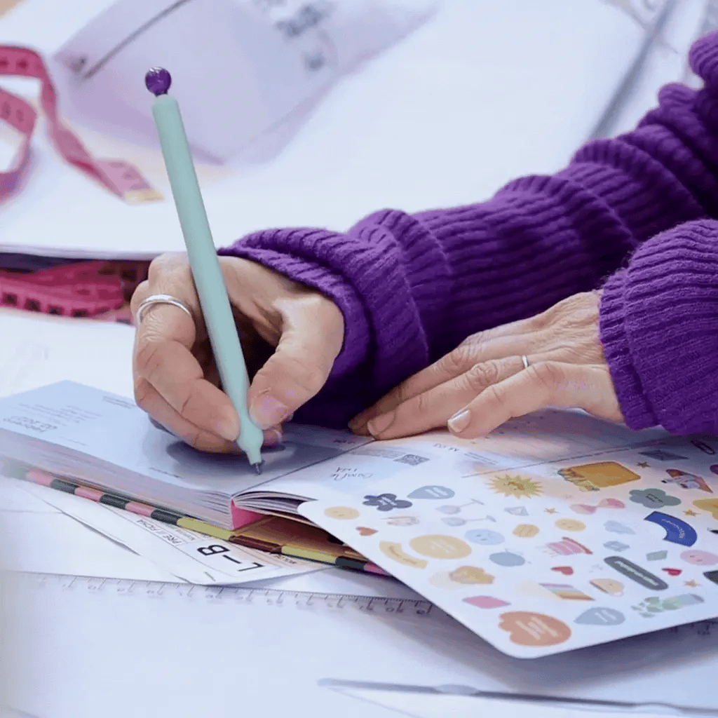 Manos escribiendo en agenda planificadora Happimess Mi Superplán 2027 con pegatinas decorativas sobre mesa de trabajo con jersey morado