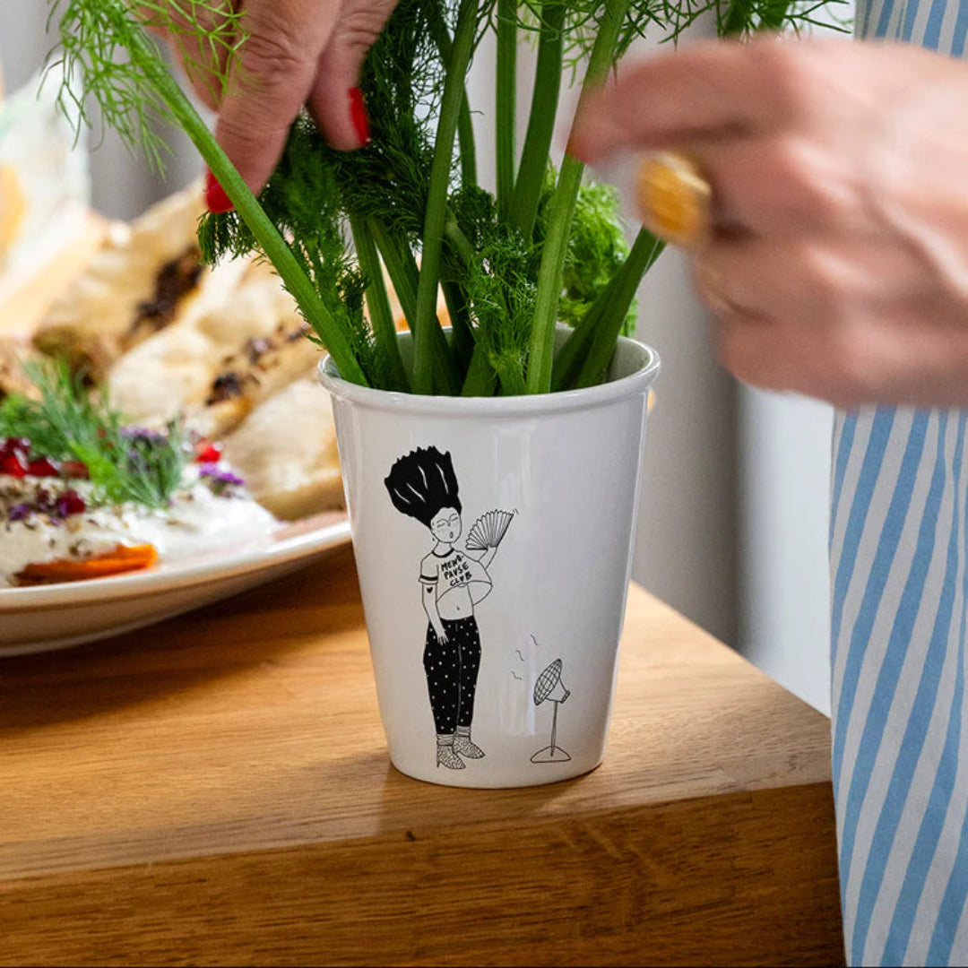 Taza en blanco y negro de una mujer abanicándose delante de un ventilador con la camiseta "Menopause Club" con ramitas de eneldo dentro manipuladas por una persona. 