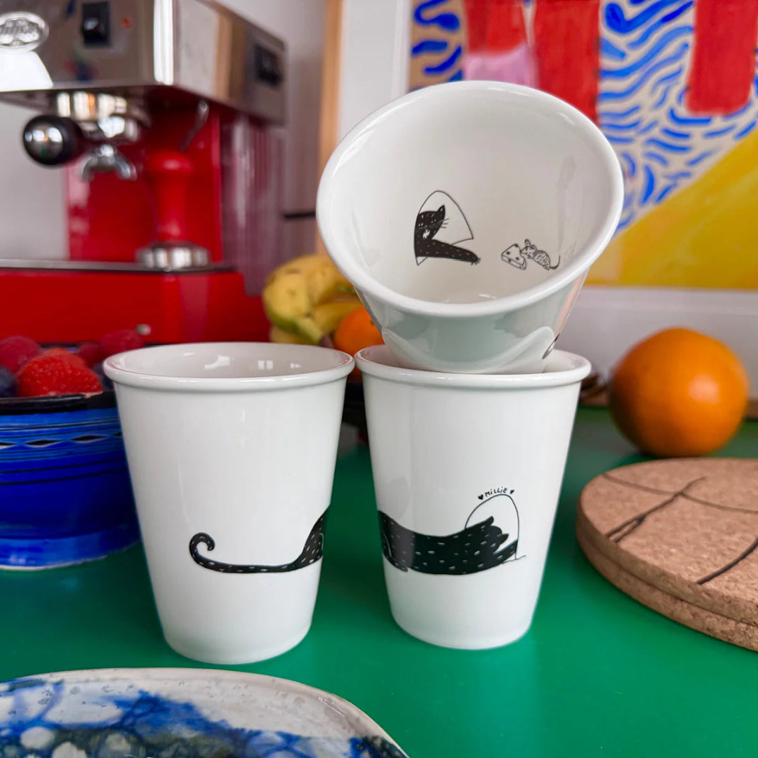 Tres tazas en blanco y negro de un gato negro, donde por fuera se ve el gato asomándose en una puerta y por dentro de la propia taza se ve la cara del gato intentando cazar un ratón con un queso. 