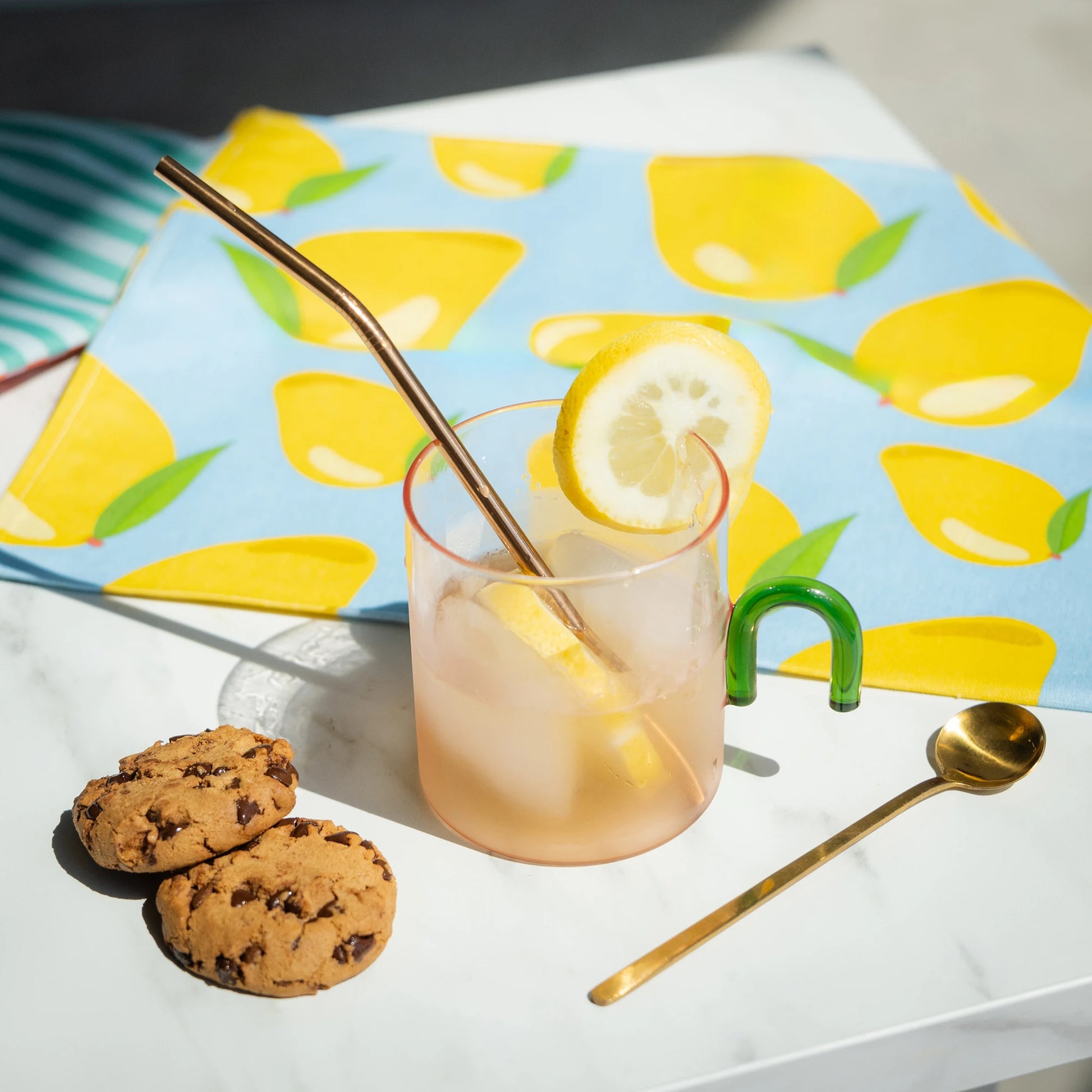 Taza de cristal rosa con asa con forma de arco verde llena de limonada junto a unas galletas con chocolate