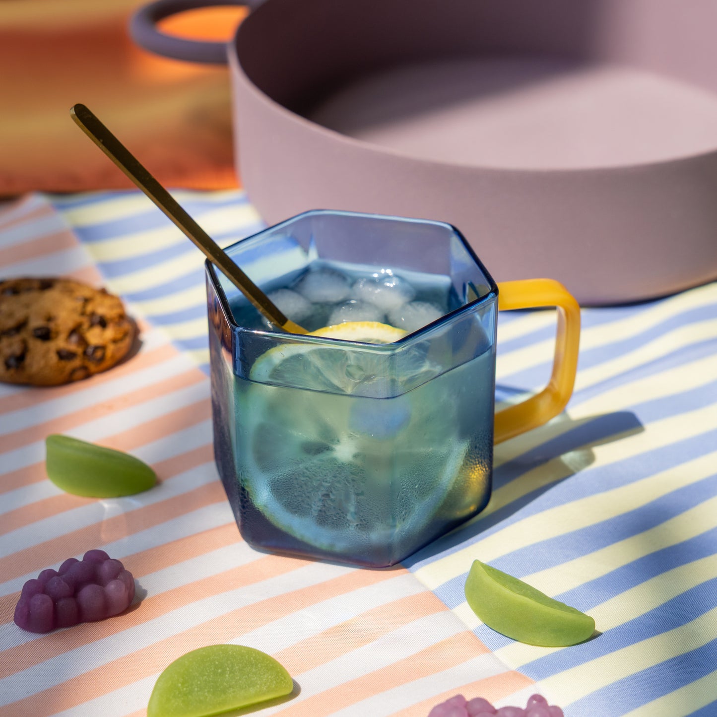 Taza hexagonal de cristal azul con mango amarillo con agua con hielo y limón sobre un mantel con galletas y gominolas