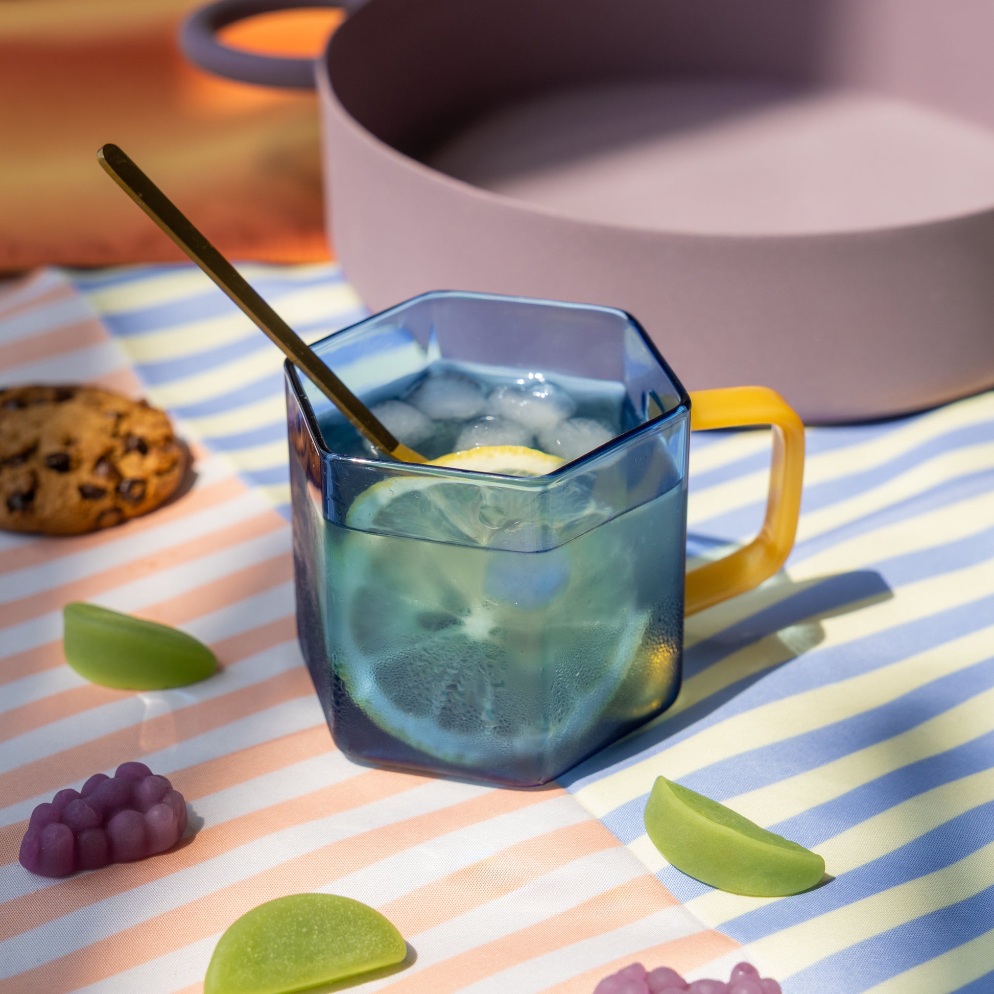 Taza hexagonal de cristal azul con mango amarillo con agua con hielo y limón sobre un mantel con galletas y gominolas