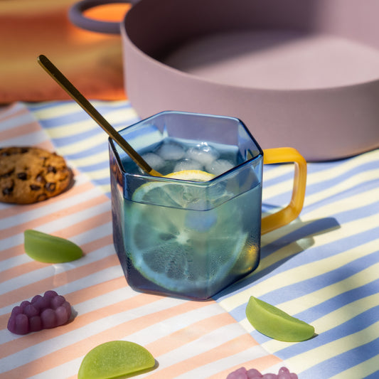 Taza hexagonal de cristal azul con mango amarillo con agua con hielo y limón sobre un mantel con galletas y gominolas