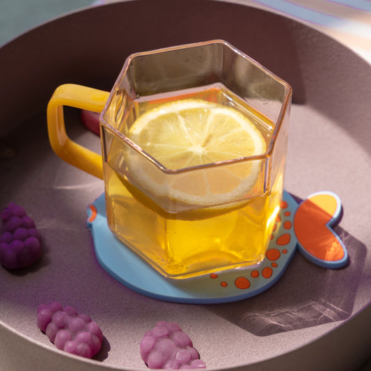 Taza hexagonal de cristal rosa con asa amarilla llena de agua con una rodaja de limón en una bandeja con chuches