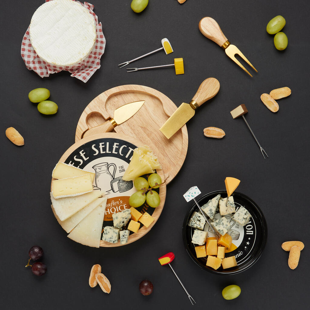 Vista aérea de tabla de quesos, uvas, frutos secos y tenedores Cheese Selection esparcidos sobre fondo oscuro junto a la lata redonda negra