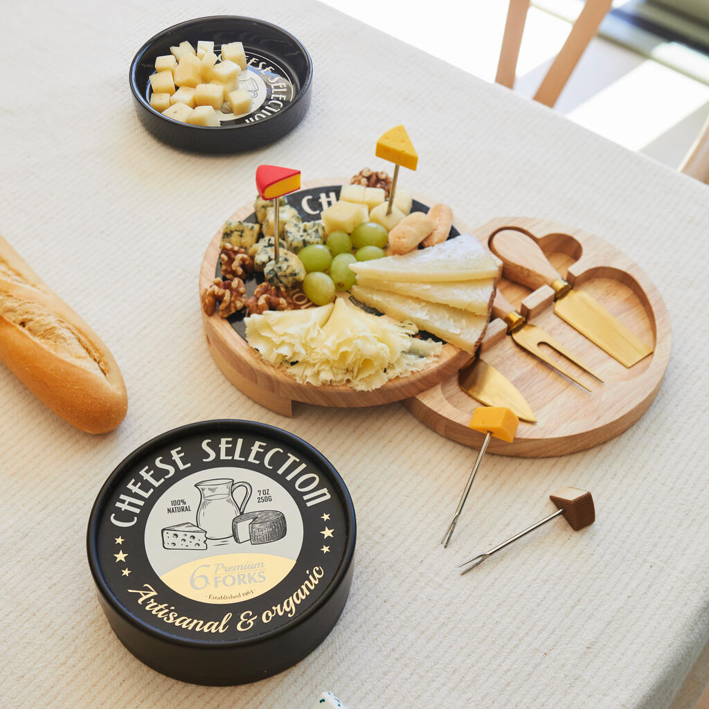 Tabla de quesos redonda de madera con tenedores Cheese Selection pinchados en distintas variedades, junto a la lata negra y una barra de pan sobre mantel blanco