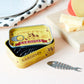 Juego de tenedores de aperitivo con forma de raspa presentados en una lata de sardinas junto a una tabla con queso