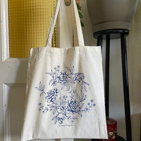 Bolsa de tela o tote bag con ilustración de flores en azul de Laura Agustí colgada en una puerta