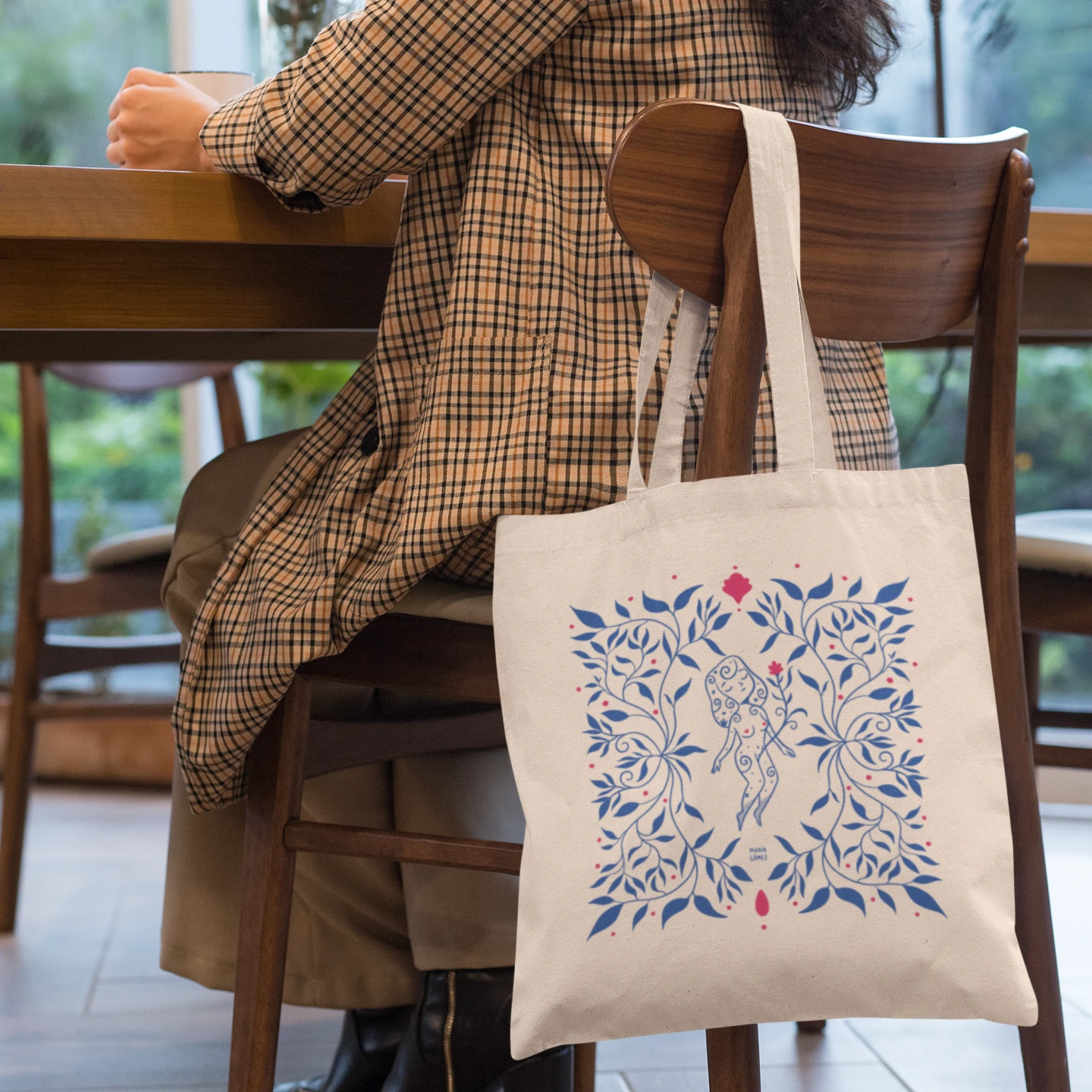 Mujer en una silla de madera tomando café con la bolsa de tela con una mujer flotando entre flores azules de María Gómez 