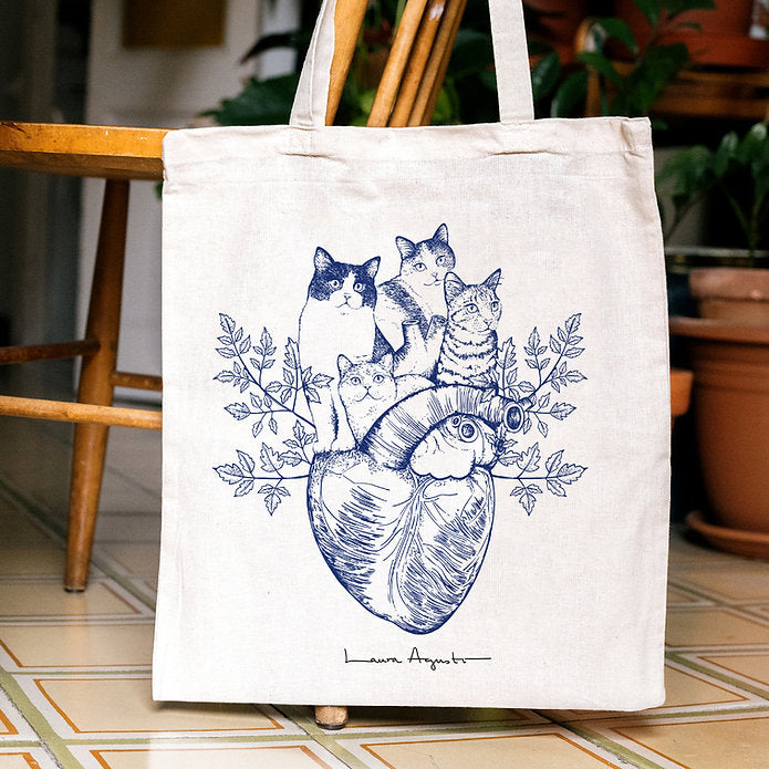 Tote bag de algodón con ilustración de cuatro gatos sobre un corazón anatómico colgada en una silla