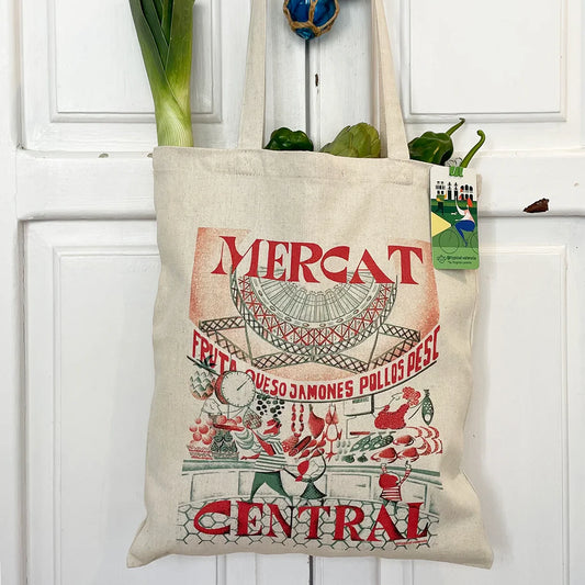 tote bag de tela con un diseño de Atypical valencia con una ilustración del mercado central de valencia y un puesto de comida con gente comprando. de la bolsa también salen frutas y verduras