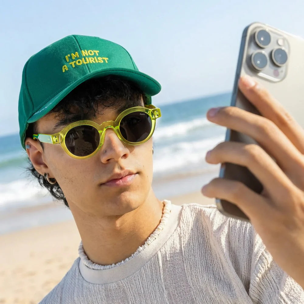 modelo posando con las gafas de sol de Fisura del modelo I'm not a tourist redondas con la parte delantera en color verde lima y los cristales en negro y las patillas en verde oscuro en la playa
