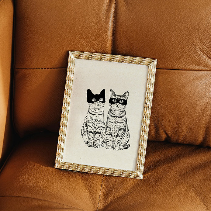 Lámina en blanco y negro de Laura Agustí de dos gatos pardos, uno con un antifaz y otro con una máscara de Batman, sobre un sofá de cuero marrón.