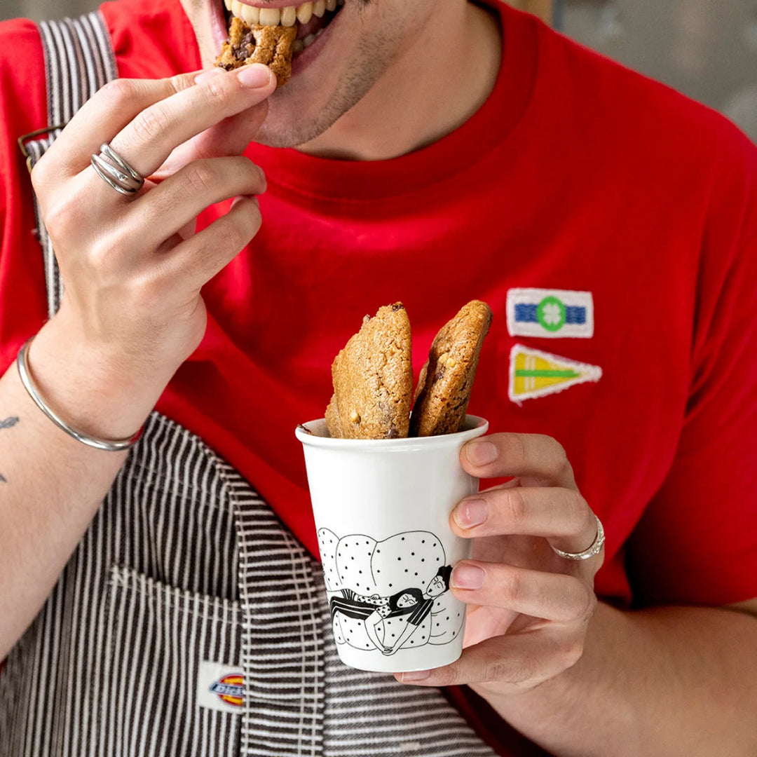 Hombre con camiseta roja comiendo galletas de un vaso de cerámica en el que aparece una pareja dormida en un sofá