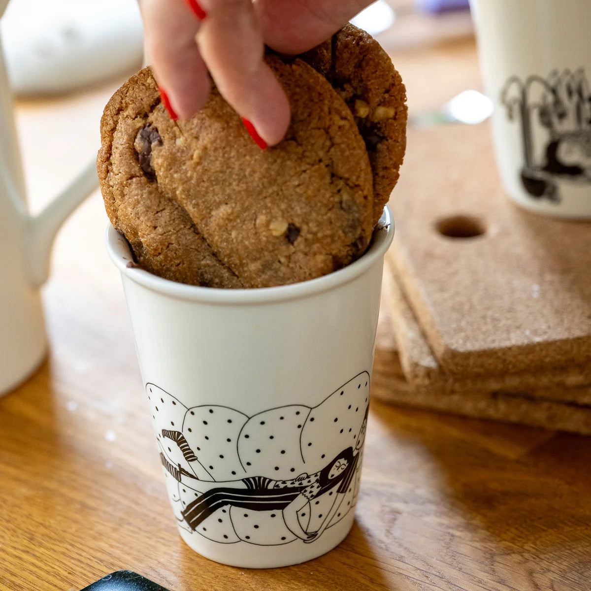 Mano cogiendo galletas del vaso de cerámica Cancelling Plans Club de Helen B con ilustración de una pareja hetero durmiendo en un sofá mullido de puntitos
