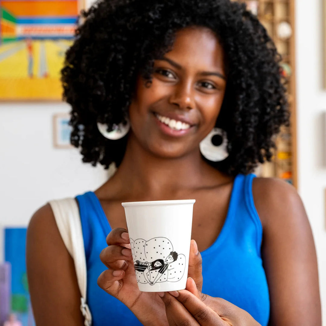 Mujer con camiseta azul sosteniendo el vaso de cerámica Cancelling Plans Club de Helen B con ilustración de una pareja hetero durmiendo en un sofá mullido de puntitos