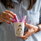 Mujer con uñas rojas y camisa de rayas azul sacando fruta morada de un vaso de cerámica con ilustración de una chica con una camiseta estampada con corazoncitos y el mensaje FUCK WHAT THEY THINK