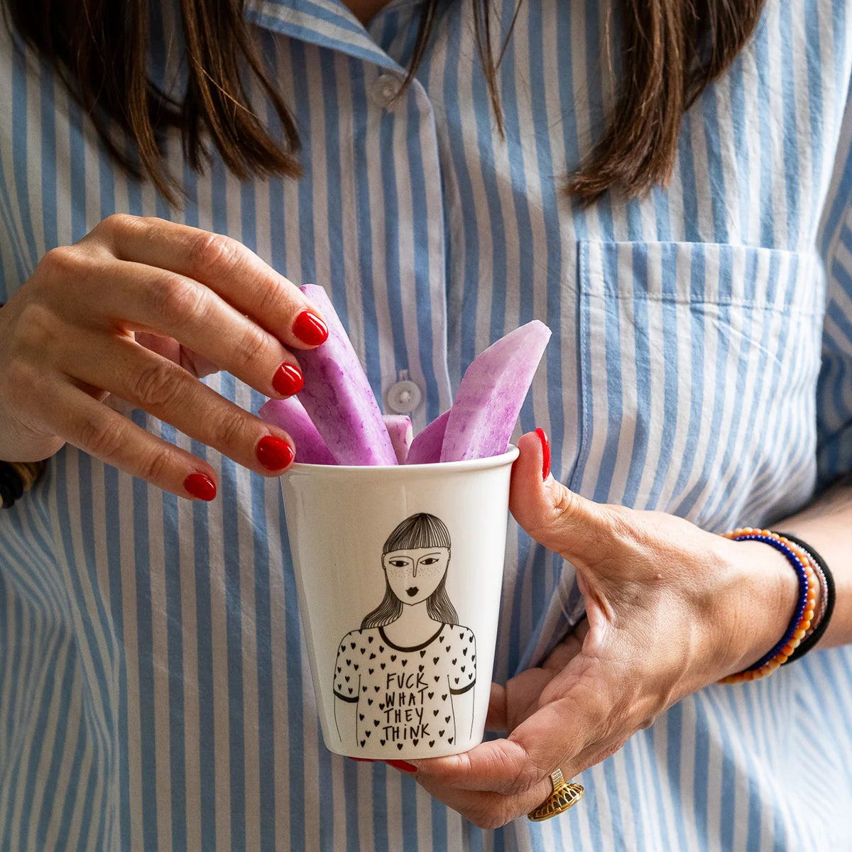 Mujer con uñas rojas y camisa de rayas azul sacando fruta morada de un vaso de cerámica con ilustración de una chica con una camiseta estampada con corazoncitos y el mensaje FUCK WHAT THEY THINK