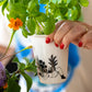 Una mujer de uñas rojas sostiene un vaso de cerámica blanca con una planta de albahaca sembrada e ilustrado por Helen B en línea negra con una chica dormida frente a unas macetas con plantas. 