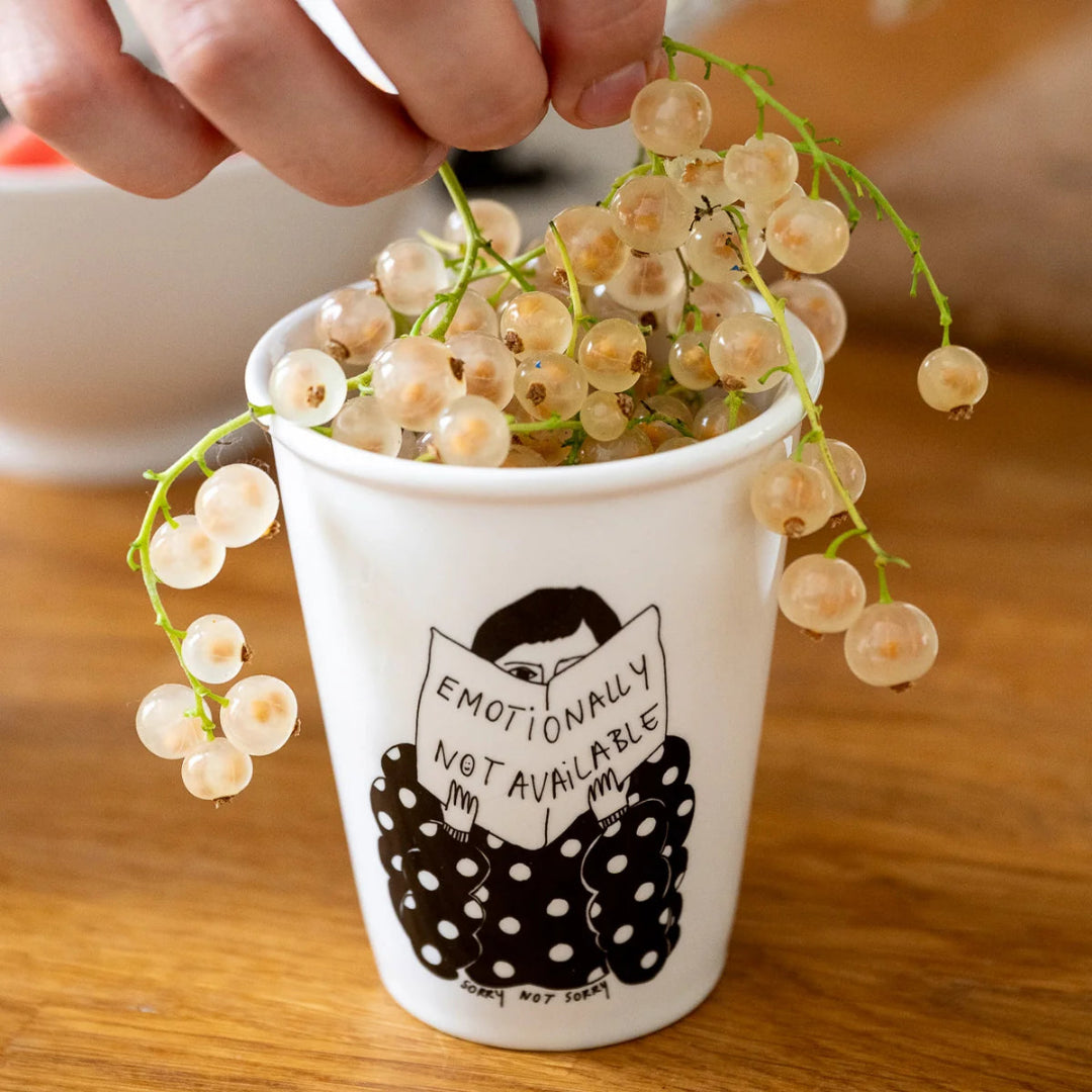 Vaso blanco Sorry Not Sorry con ilustración de figura con traje de lunares negros leyendo un libro con texto Emotionally Not Available, con uvas blancas dentro sobre superficie de madera