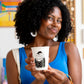 Mujer sonriendo con camiseta azul sosteniendo el vaso Sorry Not Sorry con ilustración de figura leyendo entre cuadros coloridos al fondo