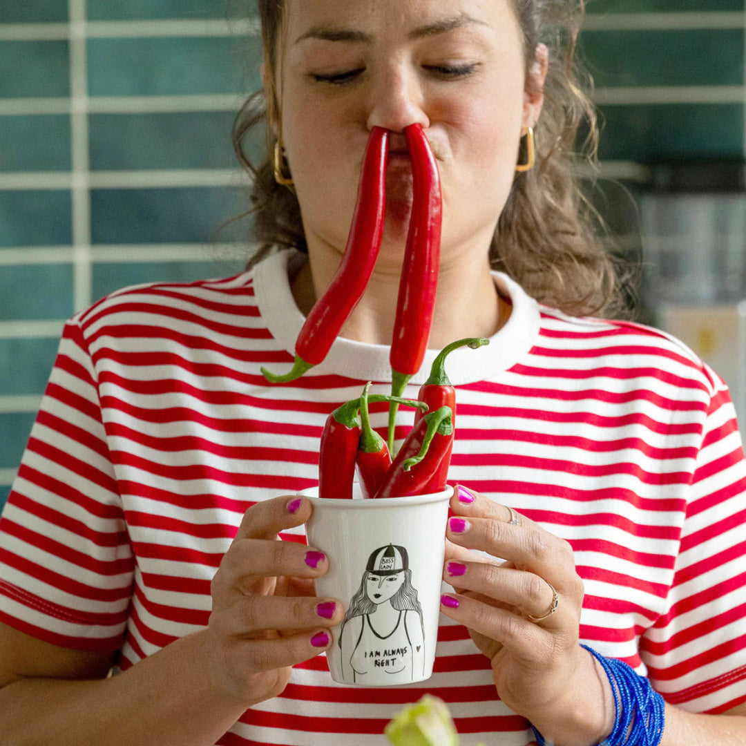 Mujer con camiseta de rayas rojas y guindillas en las narices sujetando el baso de cerámica blanco y negro llena de chillies con ilustración de una mujer que lleva una gorra en la que pone "Boss lady" y una camiseta en la que pone "I am always right" 